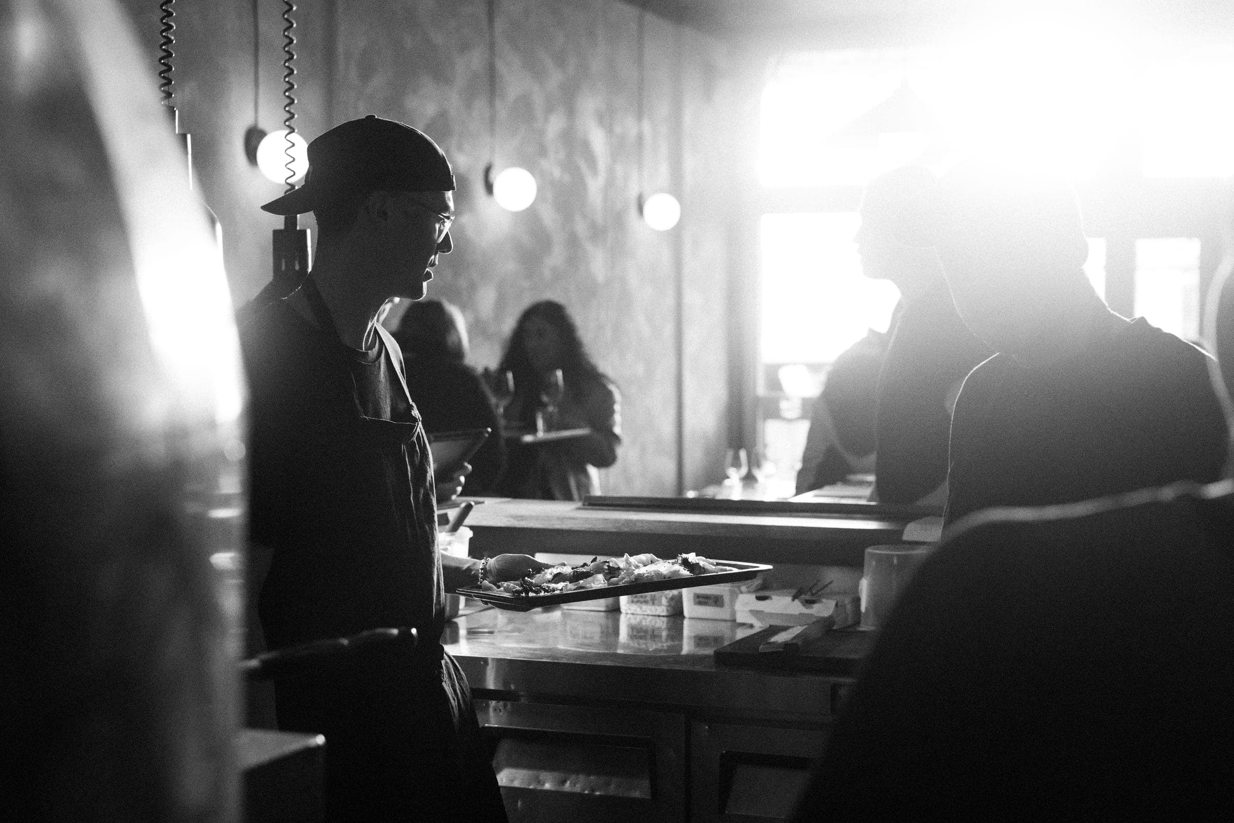 Black and white shot of a cook carrying a tray of plates past a busy bar counter