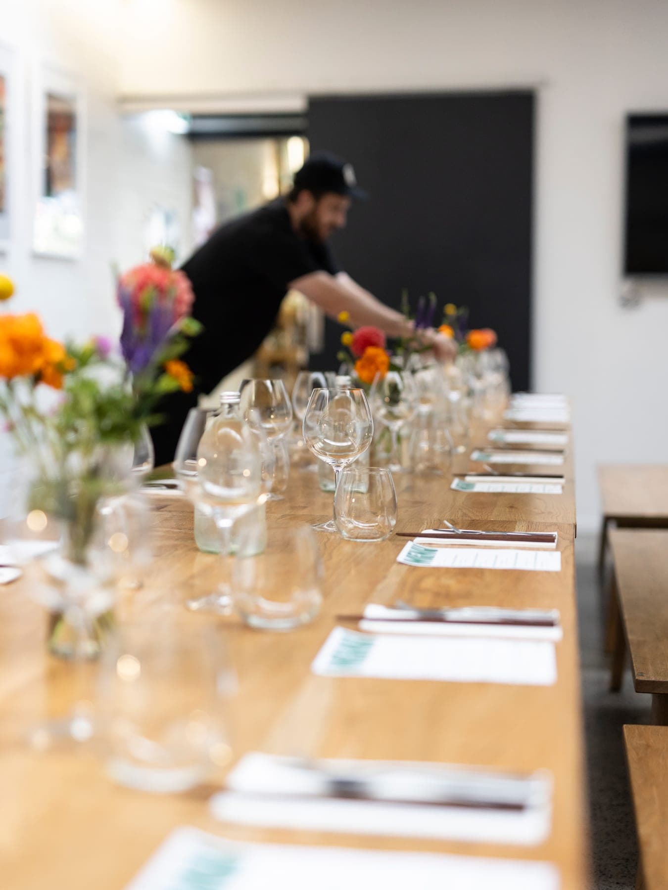 Long table set with glassware and flowers while a chef sets up in the background