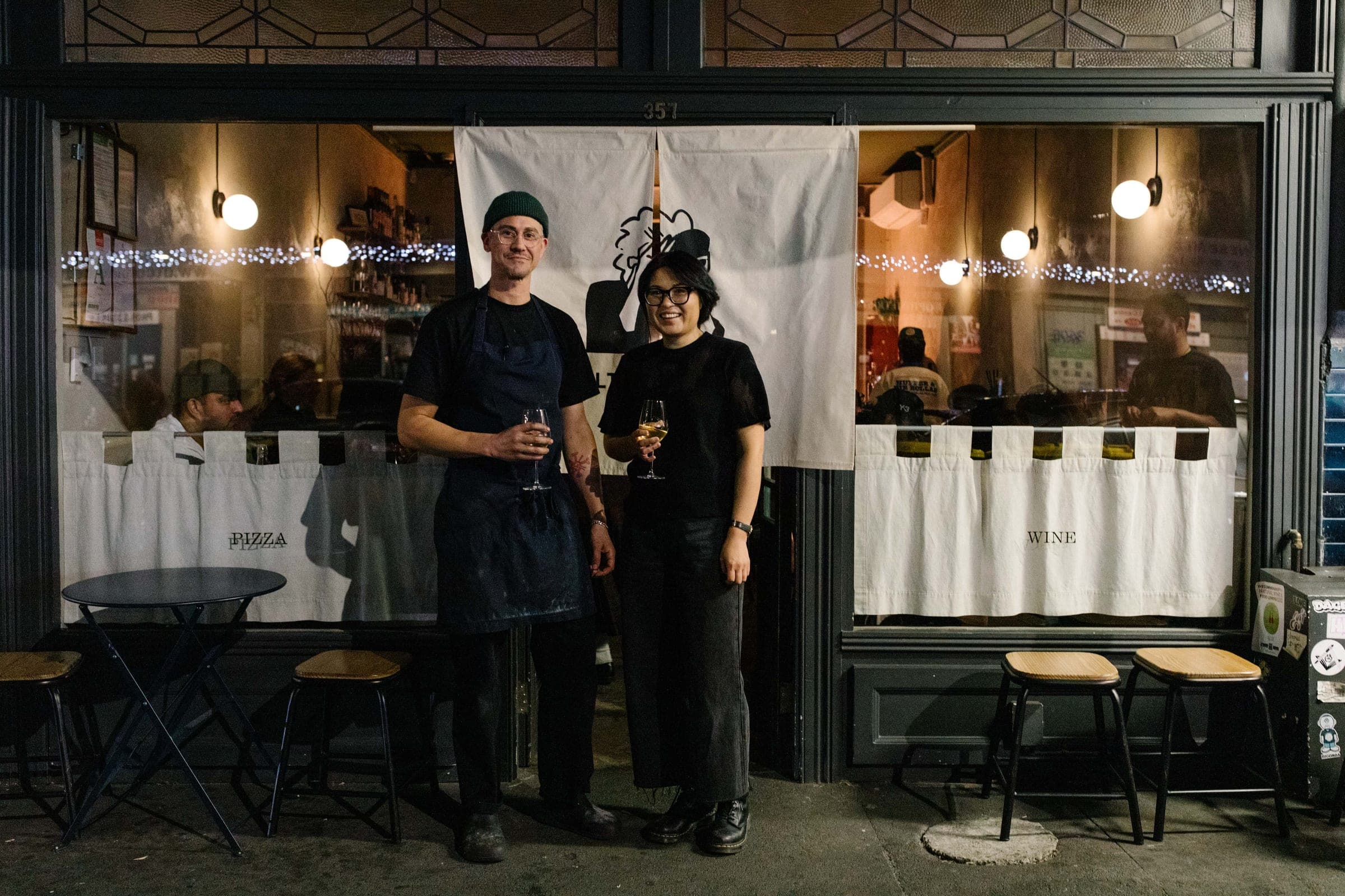 Two people in black holding glasses of wine outside a small bar at night, under a white noren curtain, with stools either side