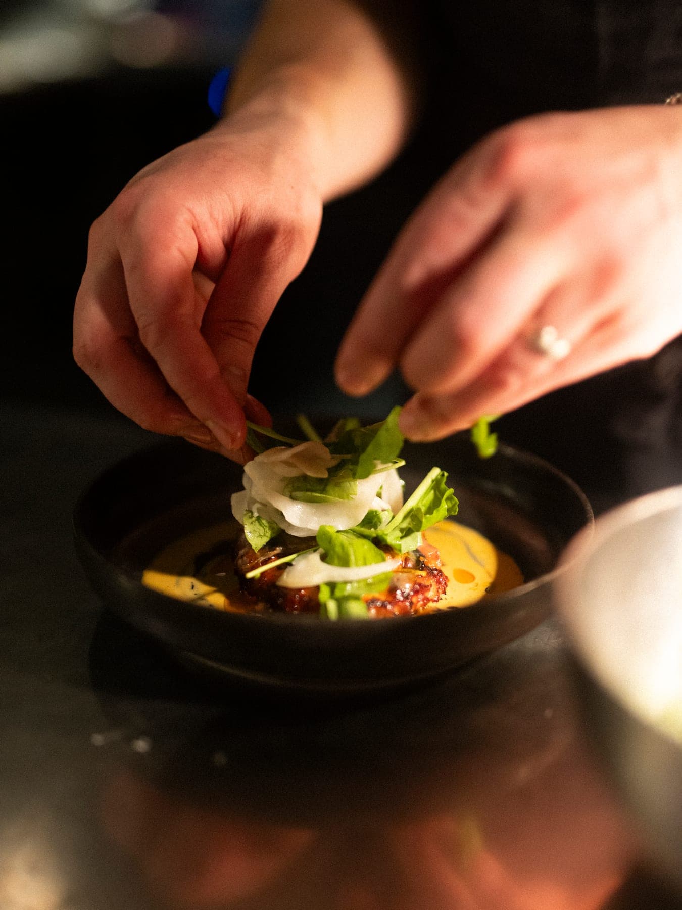 Hands finishing a dish with herbs and shaved vegetables in a black bowl at a dark pass